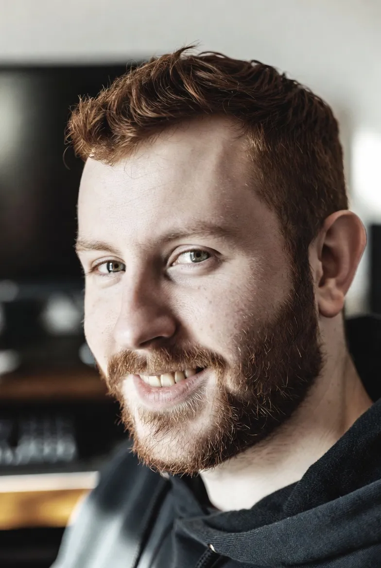A portrait of Alistair, the community strategist, styled with a rugged and weathered look. He is surrounded by atmospheric smoke and soft red glow, evoking a sense of brotherhood and leadership. The black and white portrait is grainy and tactile, capturing a professional yet approachable character who builds strong digital clans and communities.